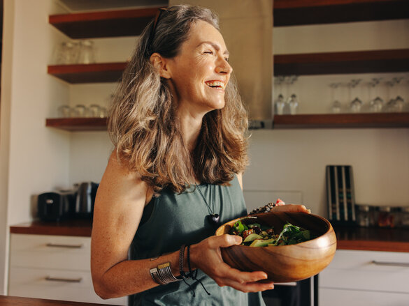 Aging woman smiling happily while holding a buddha bowl in her kitchen. Happy senior woman serving herself a healthy vegan meal at home. Mature woman taking care of her body with a plant-based diet.