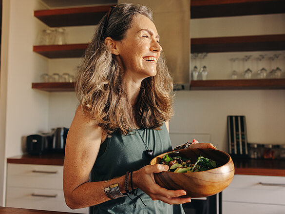 Aging woman smiling happily while holding a buddha bowl in her kitchen. Happy senior woman serving herself a healthy vegan meal at home. Mature woman taking care of her body with a plant-based diet.
