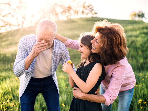Senior couple with granddaughter outside in spring nature, having fun. An old man sneezing after being tickled with a flower.